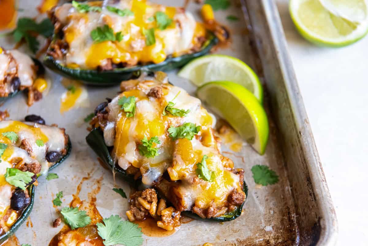 baked stuffed poblano pepper halves filled with ground beef, rice, black beans, and corn, topped with melted cheese and garnished with cilantro, with lime wedges on a sheet pan.