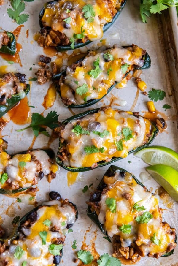 Sheet pan of baked stuffed poblano peppers filled with ground beef, rice, black beans, and corn, topped with melted cheese and garnished with cilantro and lime wedges.