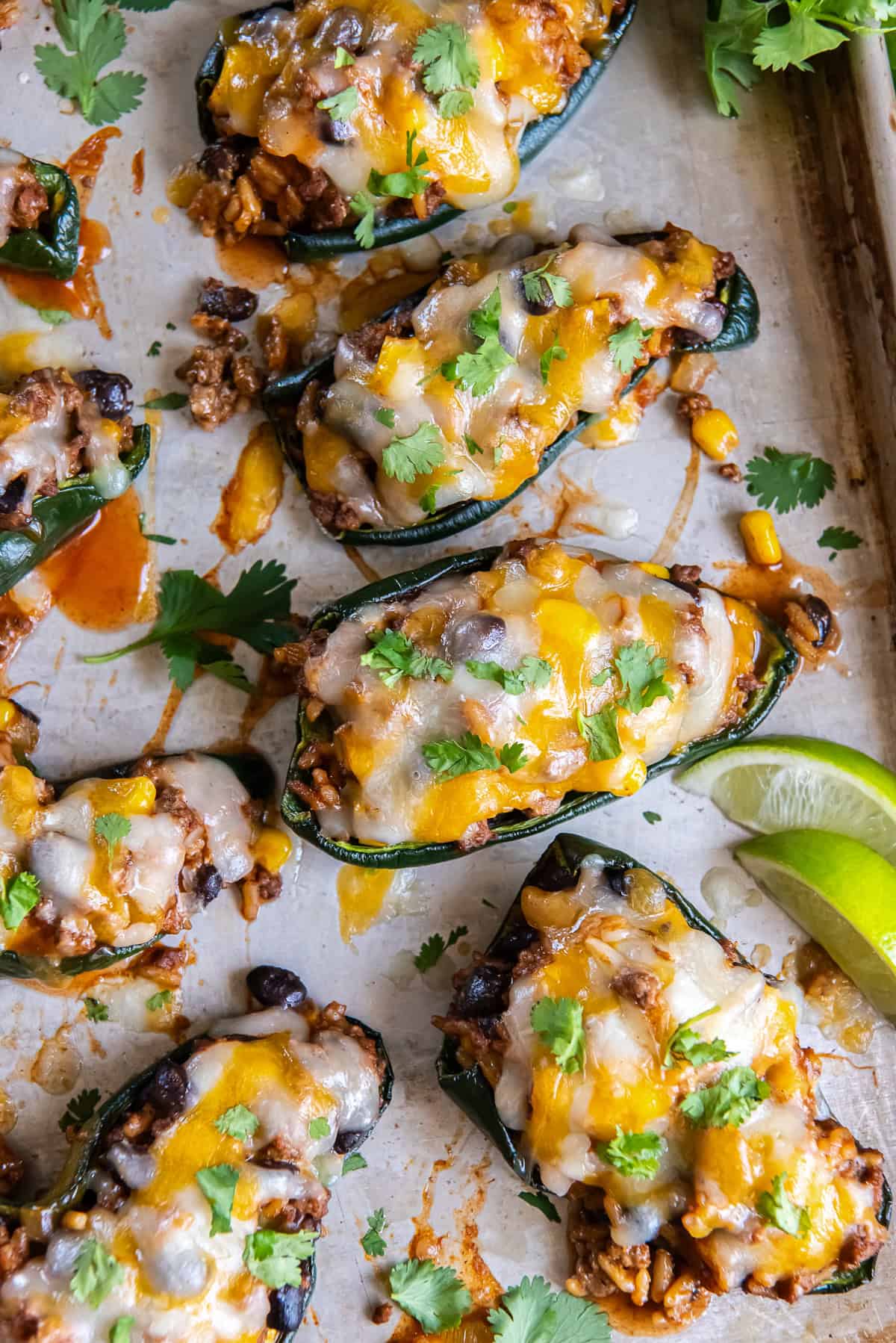 Sheet pan of baked stuffed poblano peppers filled with ground beef, rice, black beans, and corn, topped with melted cheese and garnished with cilantro and lime wedges.