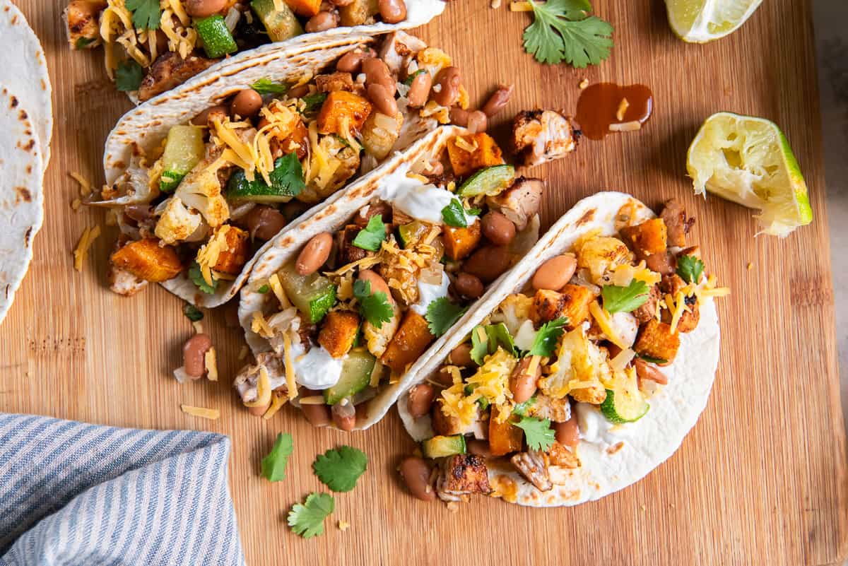 Roasted chicken and vegetable tacos topped with pinto beans, shredded cheddar cheese, cilantro, and sour cream on charred tortillas over a wooden board.