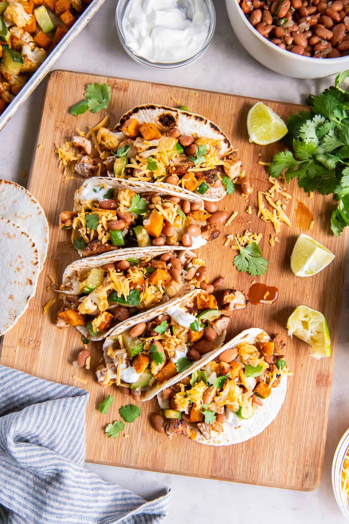 Chicken tacos with roasted sweet potatoes, cauliflower, and zucchini topped with pinto beans, shredded cheese, sour cream, and fresh cilantro on a wooden board.