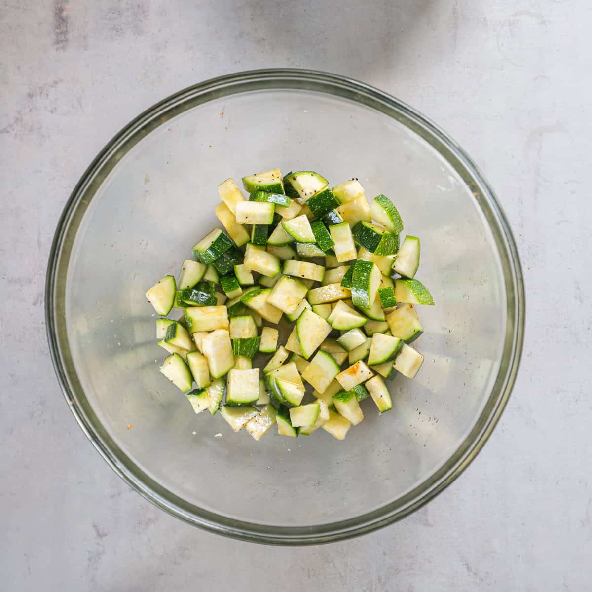 Chopped zucchini tossed with olive oil, salt, and pepper in a glass bowl before roasting.