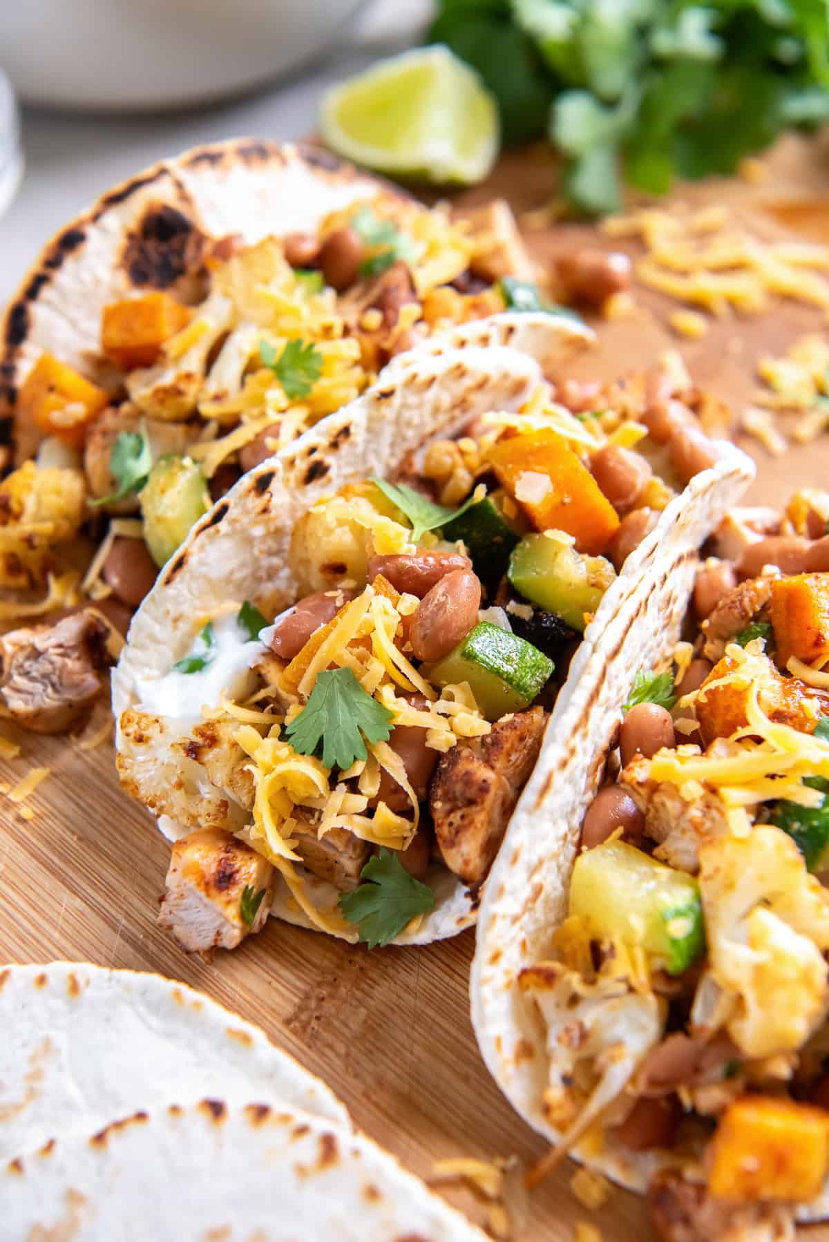 A roasted chicken and vegetable tacos topped with pinto beans, shredded cheddar cheese, cilantro, and sour cream on charred tortillas over a wooden board.