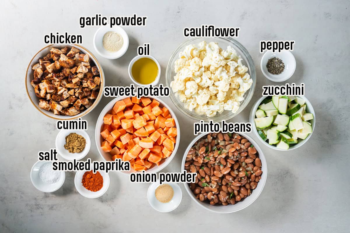 Ingredients for chicken and roasted veggie tacos including chopped chicken, sweet potato, cauliflower, zucchini, pinto beans, olive oil, cumin, smoked paprika, garlic powder, onion powder, salt, and pepper in bowls on a light background.
