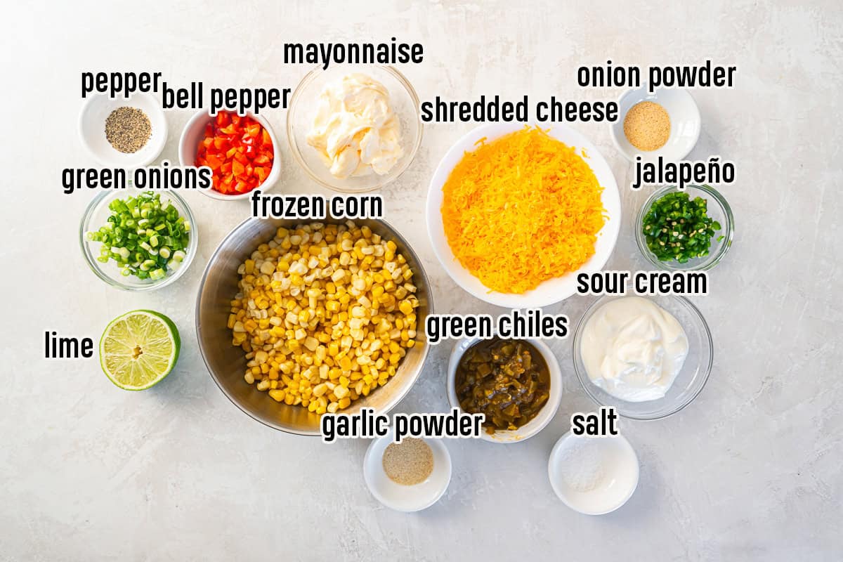 Ingredients for cold corn dip including frozen corn, cheddar cheese, sour cream, mayonnaise, green chiles, jalapeño, green onions, red bell pepper, lime, and seasonings.