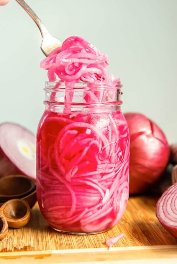 Fork lifting quick pickled red onions from a mason jar with fresh red onions in the background.