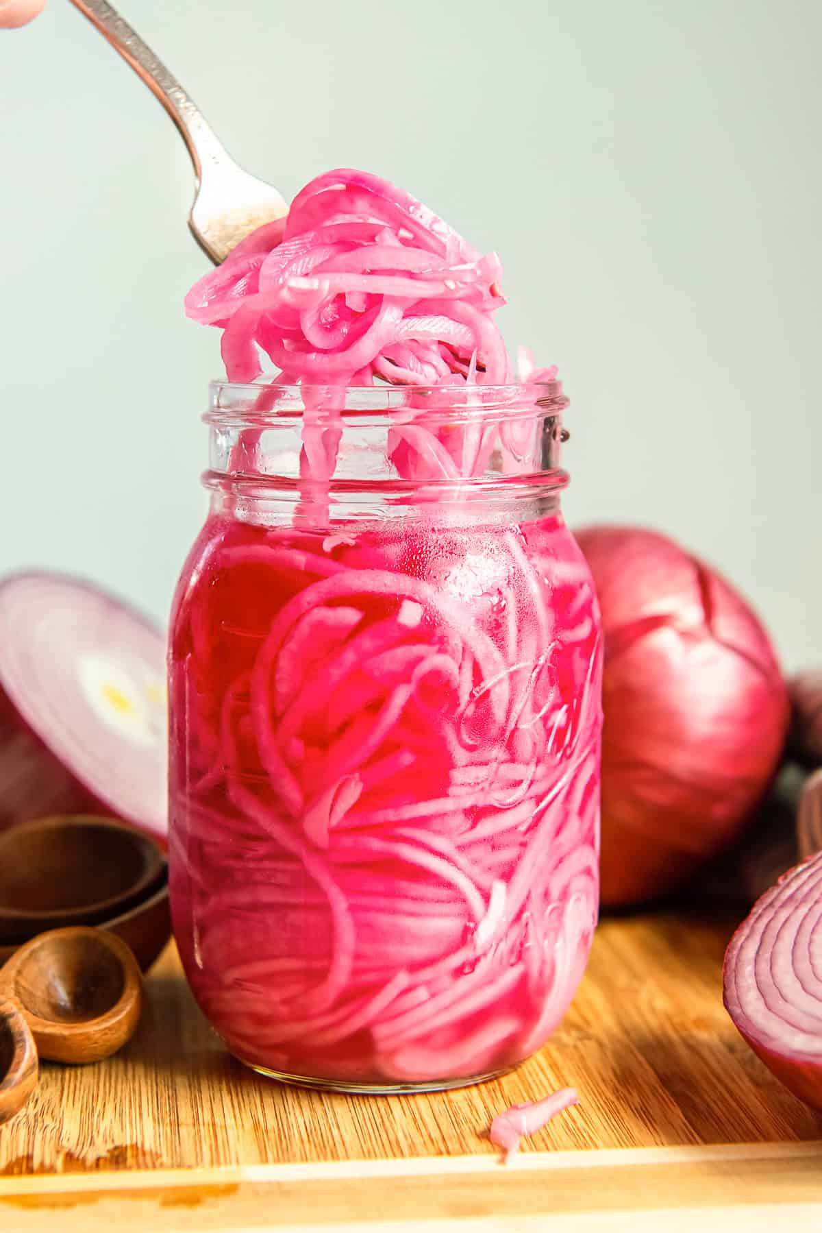 Fork lifting quick pickled red onions from a mason jar with fresh red onions in the background.
