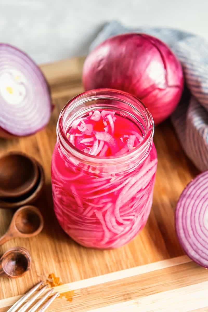 Jar of quick pickled red onions on a wooden board with fresh red onions in the background.