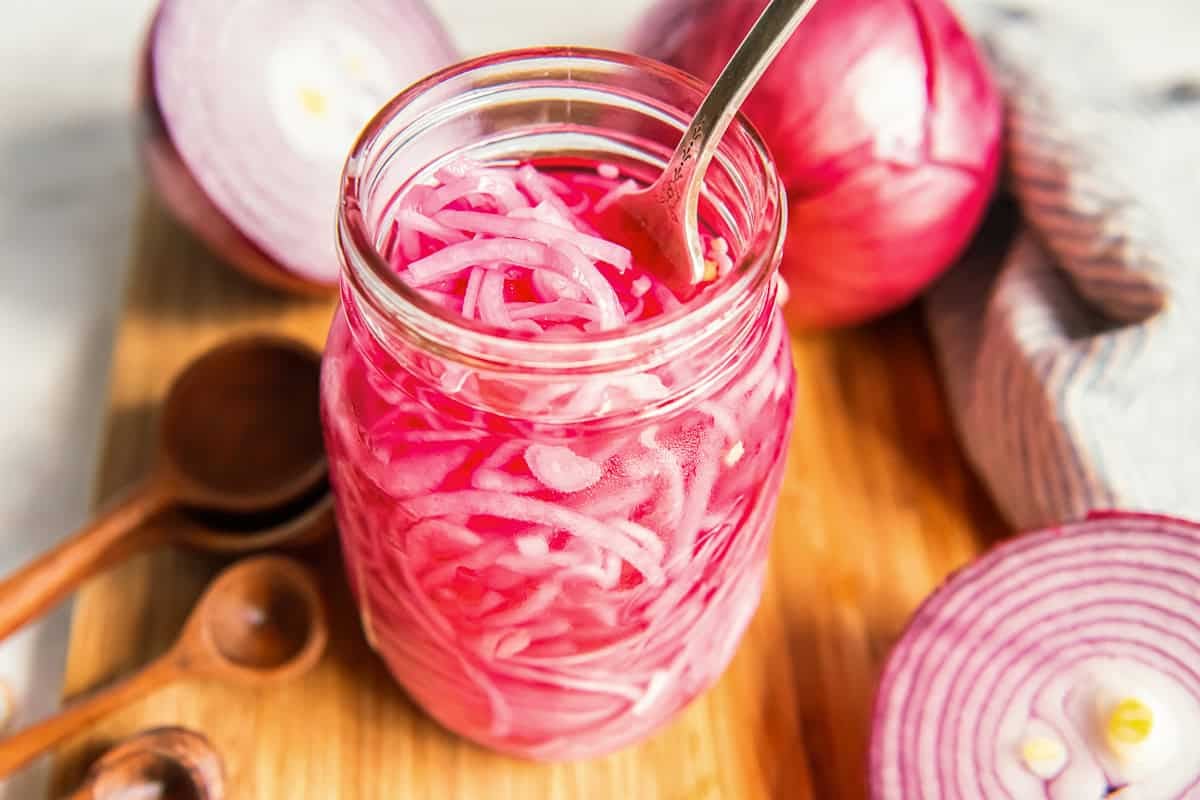 Jar of quick pickled red onions with a spoon, surrounded by fresh red onions on a wooden board.
