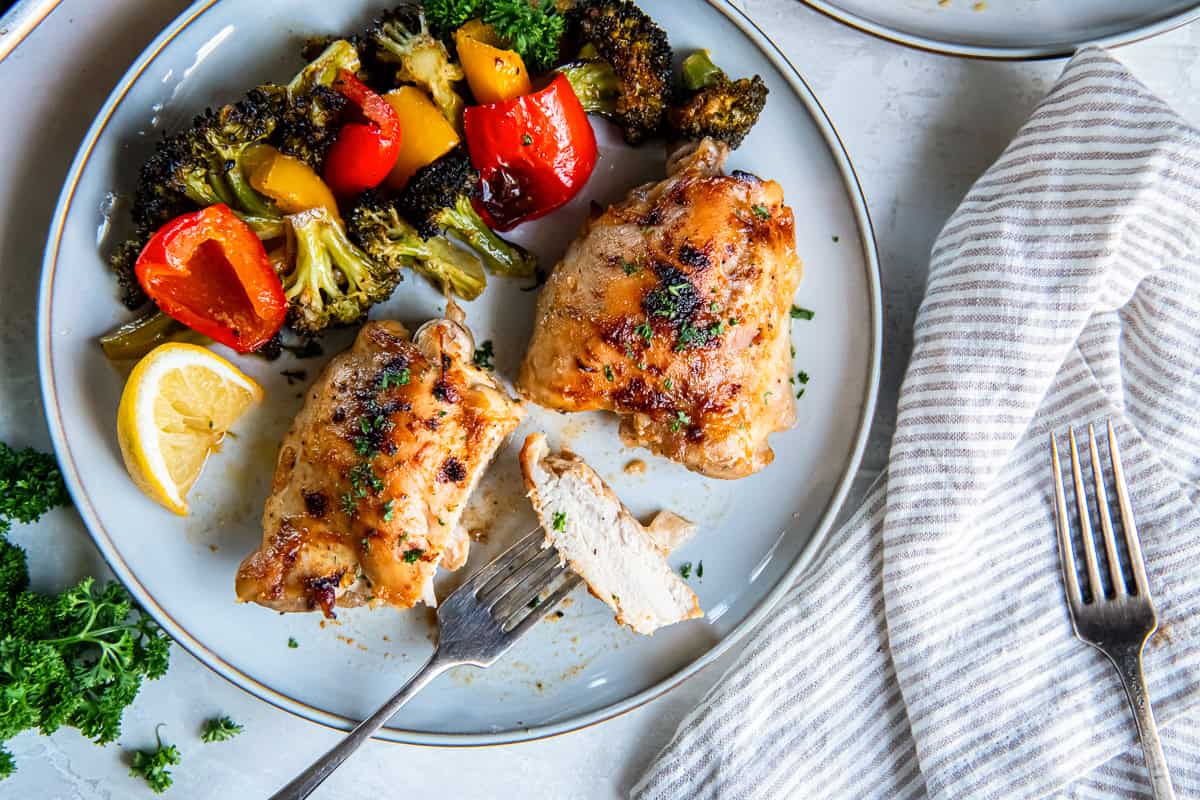 Plated honey mustard chicken thighs with roasted broccoli and red and yellow bell peppers on a gray dinner plate, with one sliced piece of chicken resting on a fork and a lemon wedge on the side.