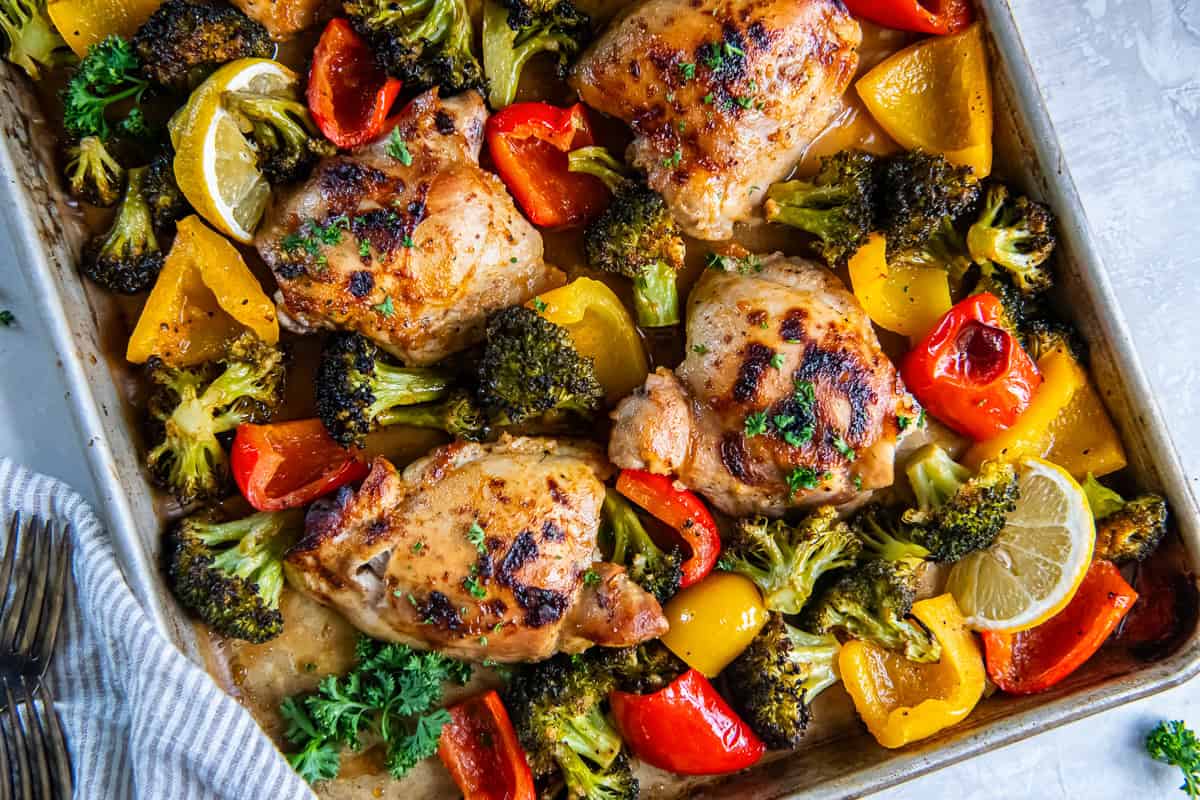 Sheet pan honey mustard chicken thighs with roasted broccoli and red and yellow bell peppers, garnished with parsley and lemon wedges.
