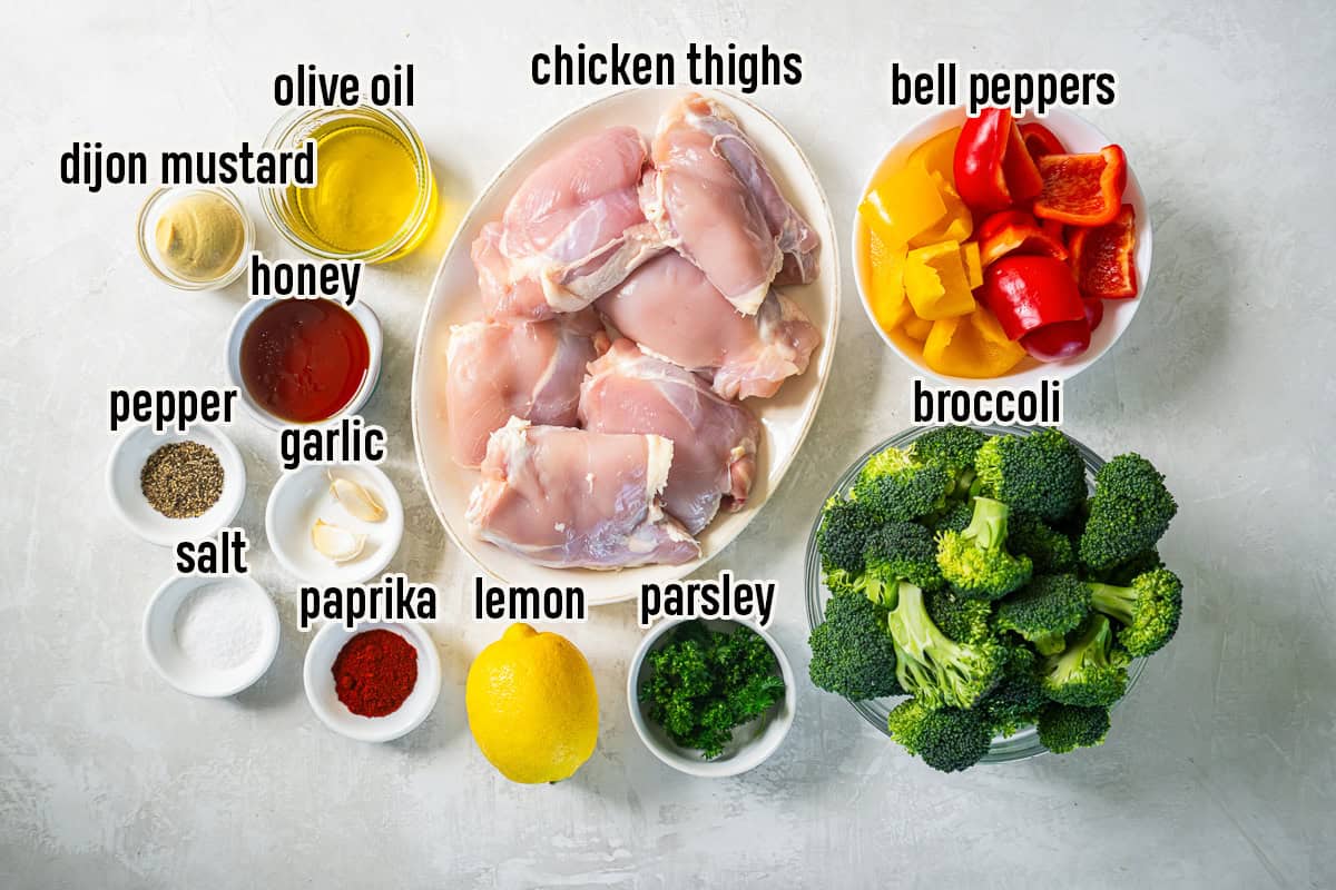 Ingredients for Sheet Pan Honey Mustard Chicken arranged on a light surface, including chicken thighs, broccoli florets, bell peppers, olive oil, Dijon mustard, honey, garlic, lemon, paprika, parsley, salt, and black pepper.