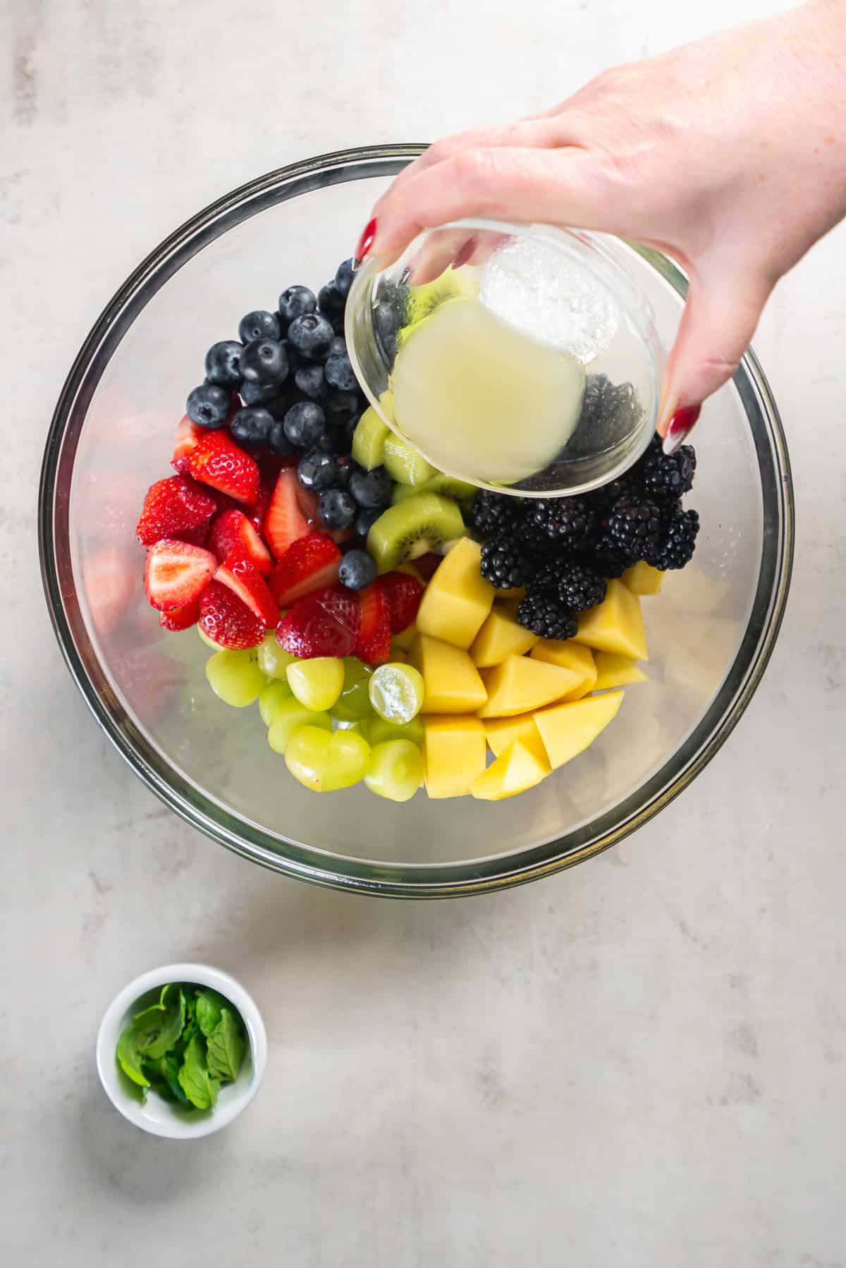 Pouring a sweet lime dressing over fresh strawberries, blueberries, blackberries, kiwi, grapes, and mango in a glass bowl for fruit salad.