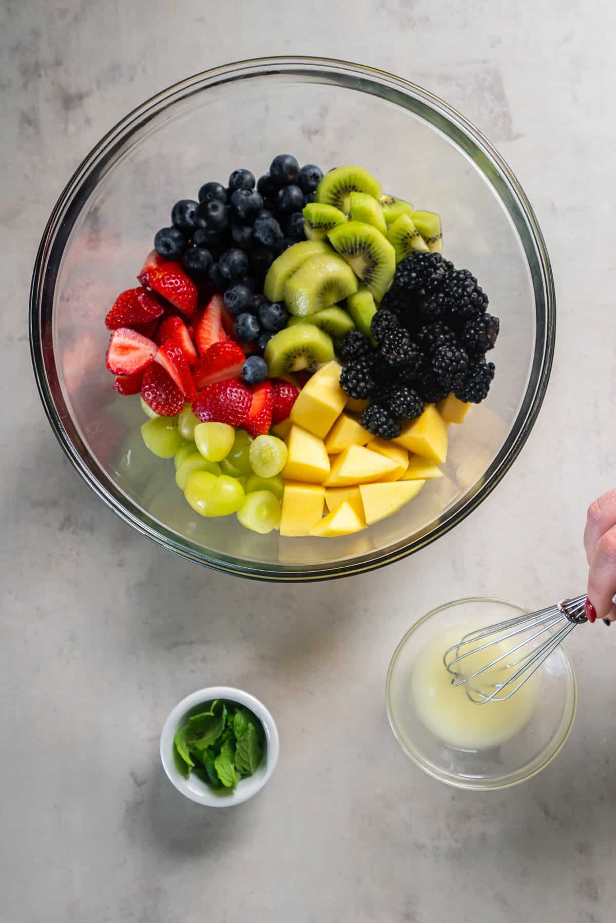 Fresh strawberries, blueberries, blackberries, kiwi, grapes, and mango in a glass bowl with a simple lime dressing being whisked nearby.