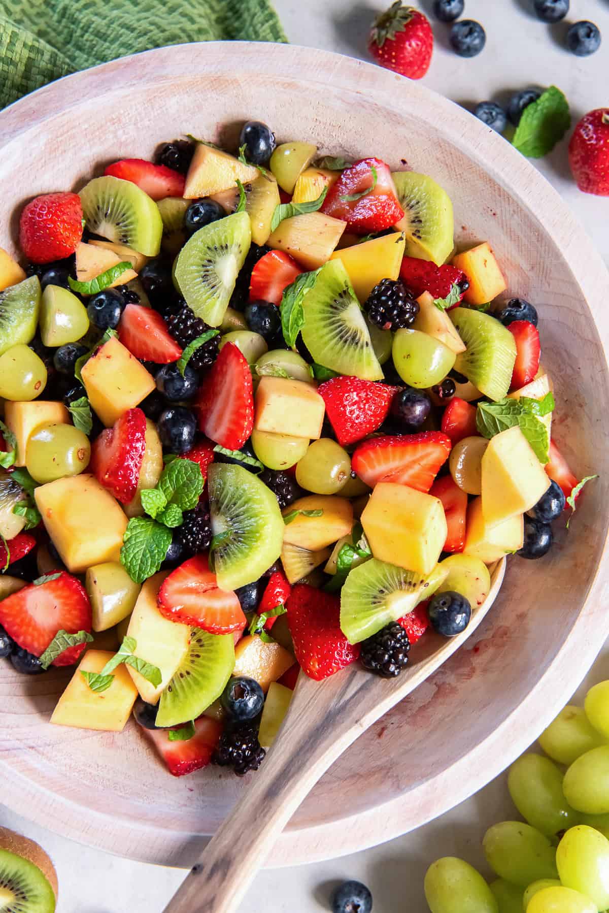 Simple fruit salad with strawberries, kiwi, grapes, mango, blueberries, blackberries, and fresh mint in a wooden bowl with a wooden serving spoon.