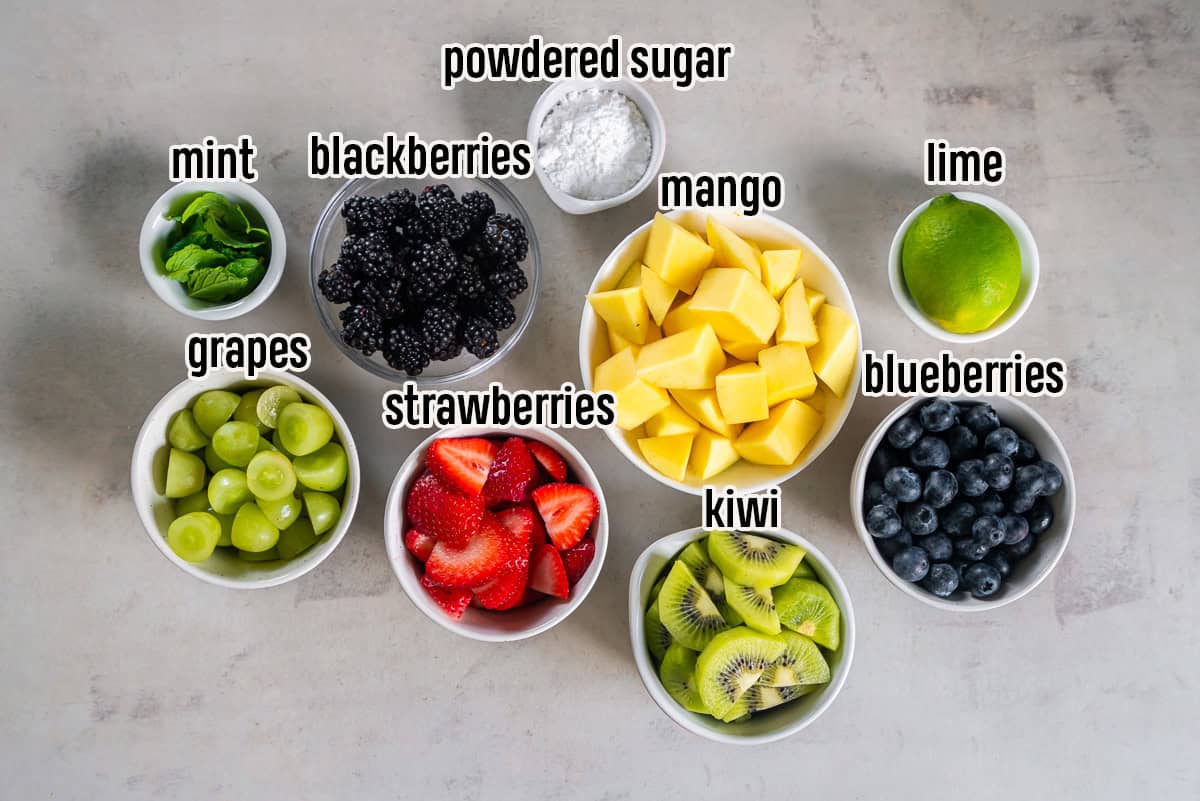 Ingredients for Simple Fruit Salad including strawberries, blueberries, blackberries, grapes, kiwi, mango, fresh mint, lime, and powdered sugar arranged in bowls on a light surface.