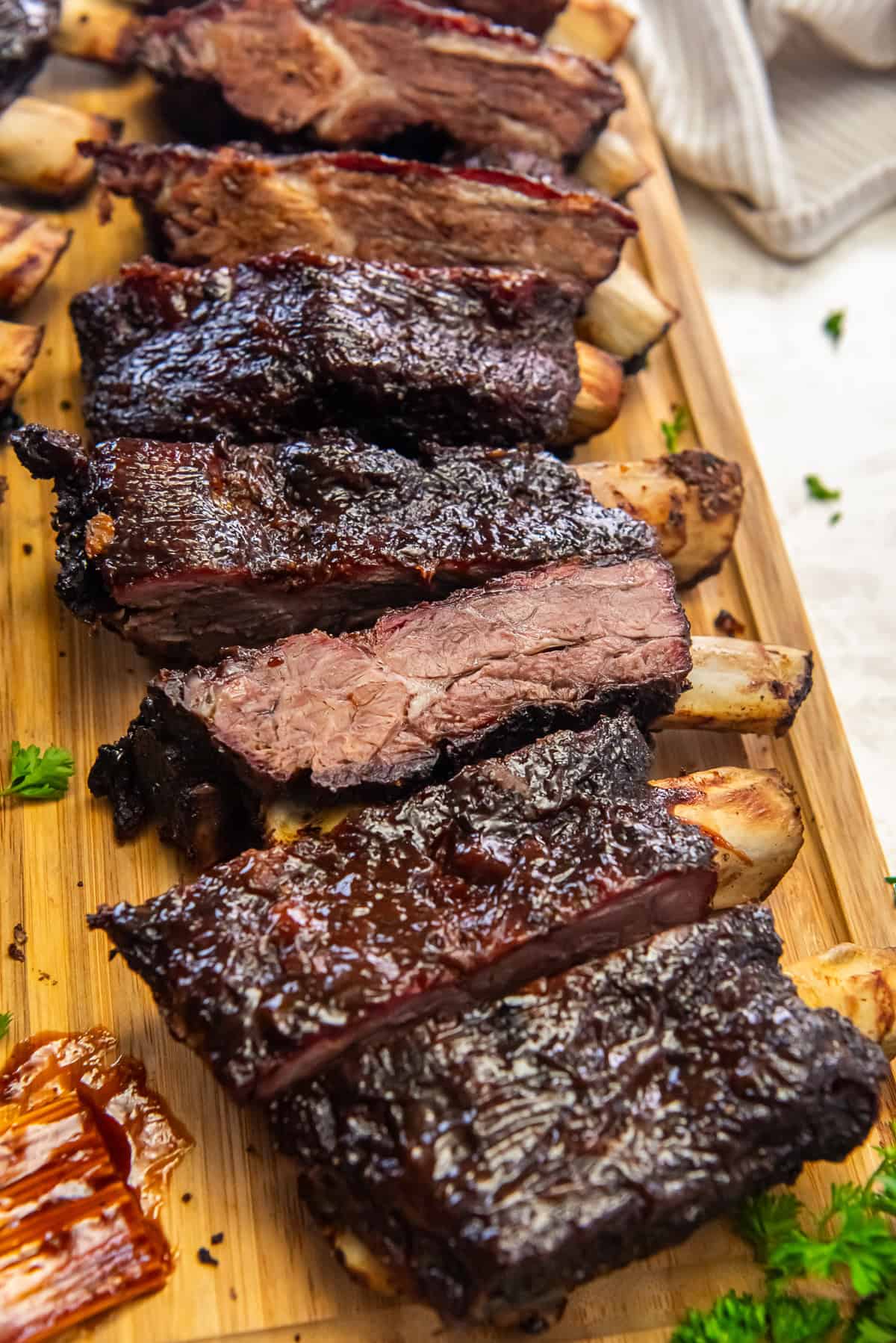 Smoked beef ribs sliced on a wooden board with a dark, caramelized bark and juicy, tender meat, served with barbecue sauce and fresh parsley.