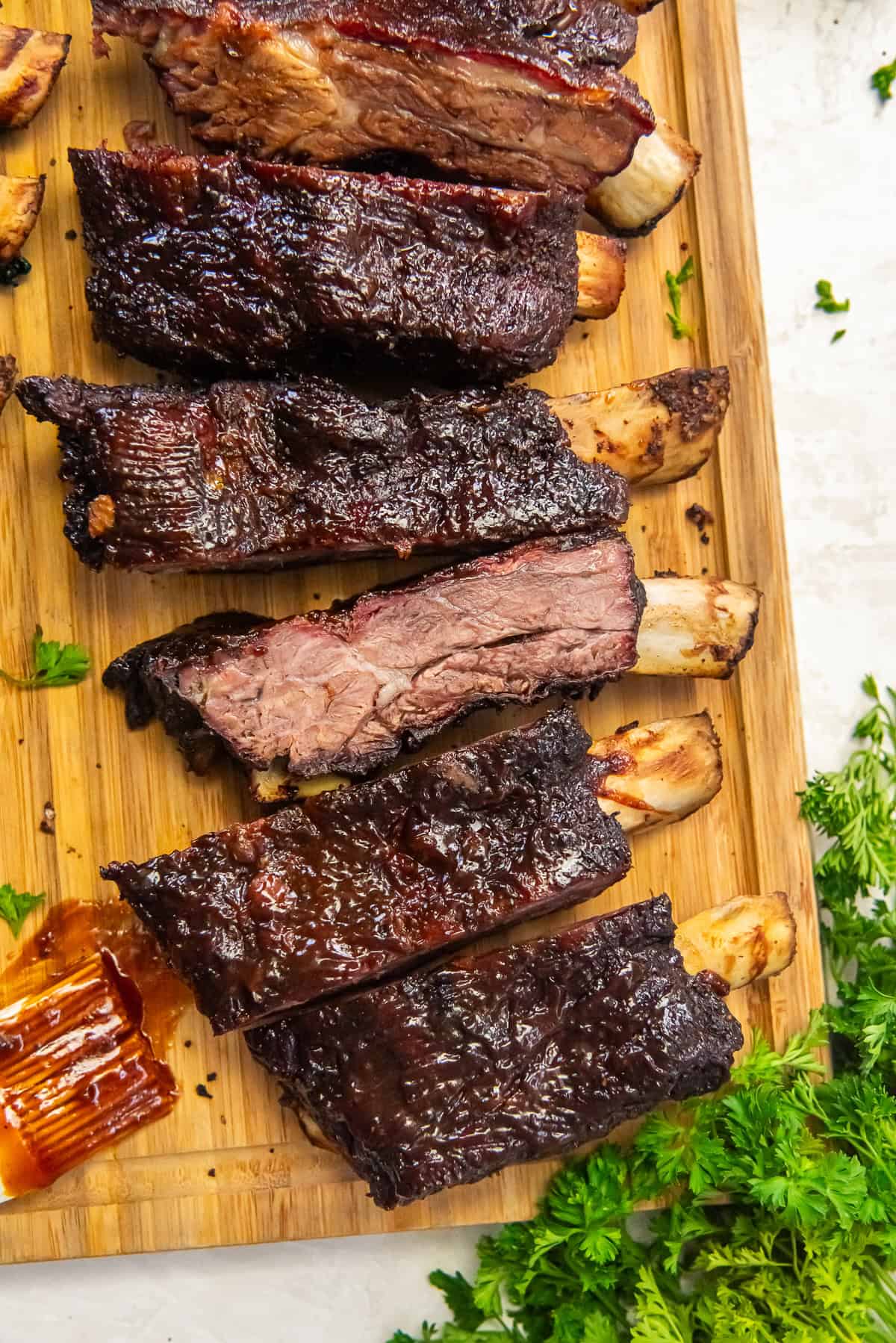 Smoked beef ribs sliced on a wooden board with a dark, caramelized bark and juicy, tender meat, served with barbecue sauce and fresh parsley.