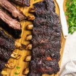 Smoked beef back ribs with a dark, flavorful bark on a wooden cutting board with sliced ribs, barbecue sauce, and fresh parsley.