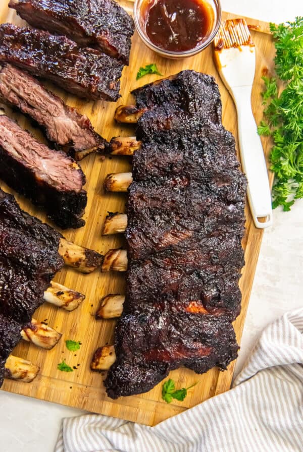 Smoked beef back ribs with a dark, flavorful bark on a wooden cutting board with sliced ribs, barbecue sauce, and fresh parsley.