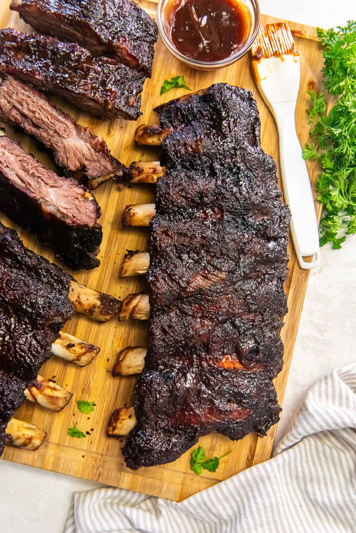 Smoked beef back ribs with a dark, flavorful bark on a wooden cutting board with sliced ribs, barbecue sauce, and fresh parsley.
