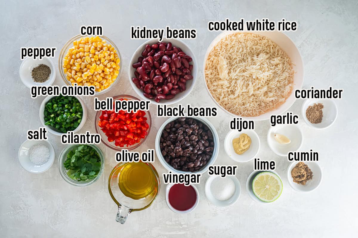 Ingredients for a rice and bean salad including cooked rice, black beans, kidney beans, corn, red bell pepper, green onions, cilantro, and dressing ingredients arranged on a countertop.