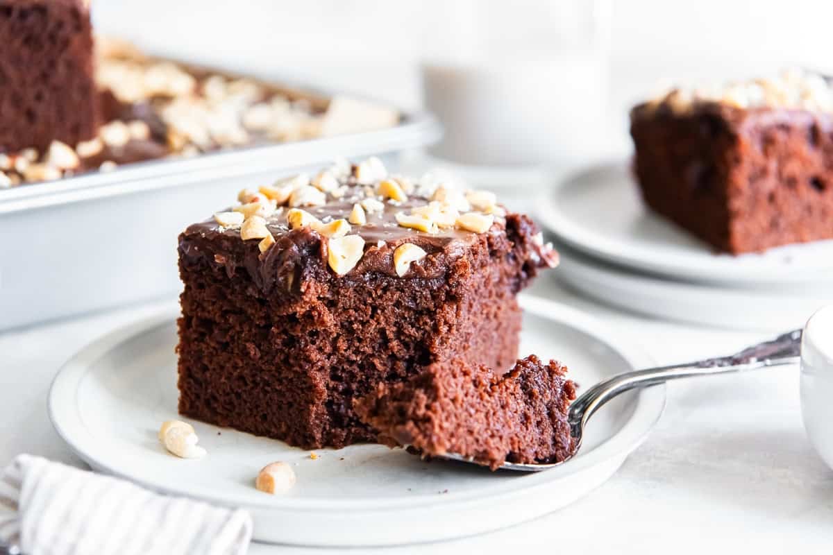 Chocolate peanut butter cake topped with rich chocolate frosting and chopped peanuts on a white plate with a bite on a spoon, with the sheet cake and a second slice blurred in the background.