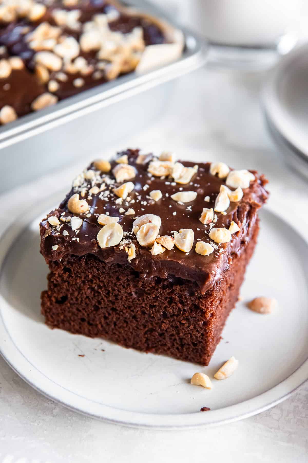 Square slice of chocolate peanut butter cake with chocolate frosting and chopped peanuts served on a white plate, with the baking pan and a glass of milk softly blurred in the background.