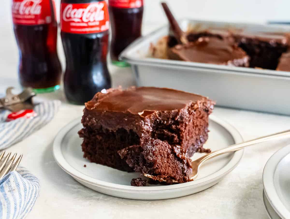 A slice of Coca-Cola chocolate cake on a small plate with a bite on a fork in the foreground. The cake is topped with a glossy chocolate frosting, with glass Coca-Cola bottles and the remaining cake in the background.