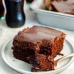 A slice of Coca-Cola chocolate cake on a small plate with a bite on a fork in the foreground. The cake is topped with a glossy chocolate frosting, with a glass Coca-Cola bottle and the remaining cake in the background.
