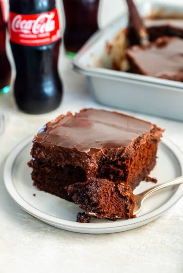 A slice of Coca-Cola chocolate cake on a small plate with a bite on a fork in the foreground. The cake is topped with a glossy chocolate frosting, with a glass Coca-Cola bottle and the remaining cake in the background.