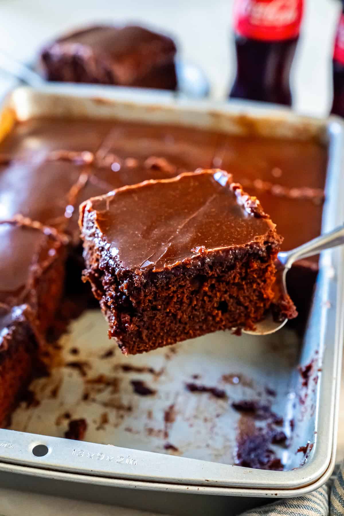 A slice of Coca-Cola chocolate cake lifted from a sheet pan on a metal serving spatula. The cake is topped with a glossy chocolate frosting, with blurred vintage Coca-Cola bottles in the background.