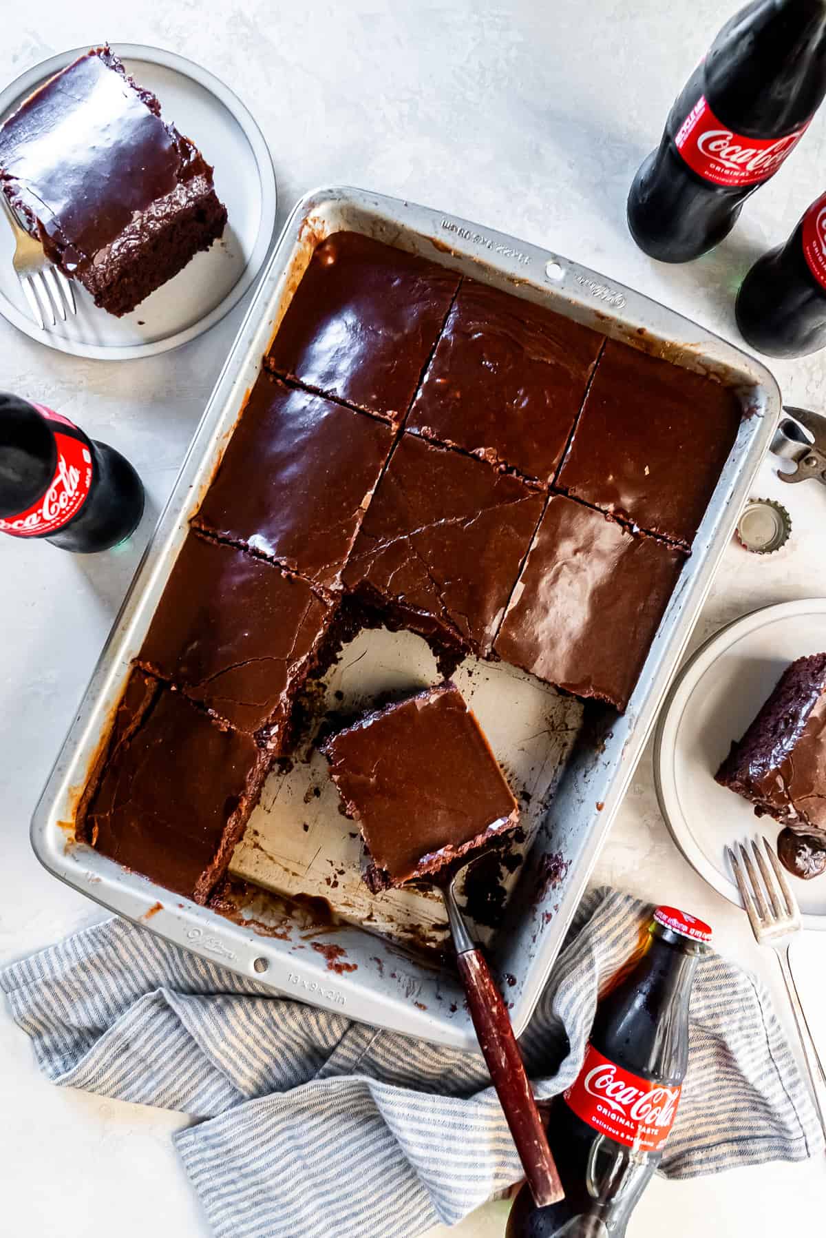 Coca-Cola sheet cake in a metal baking pan with one slice lifted on a serving spatula. The chocolate cake is topped with a glossy chocolate frosting and surrounded by vintage glass Coca-Cola bottles, plated cake slices, a bottle cap, and a striped kitchen towel.