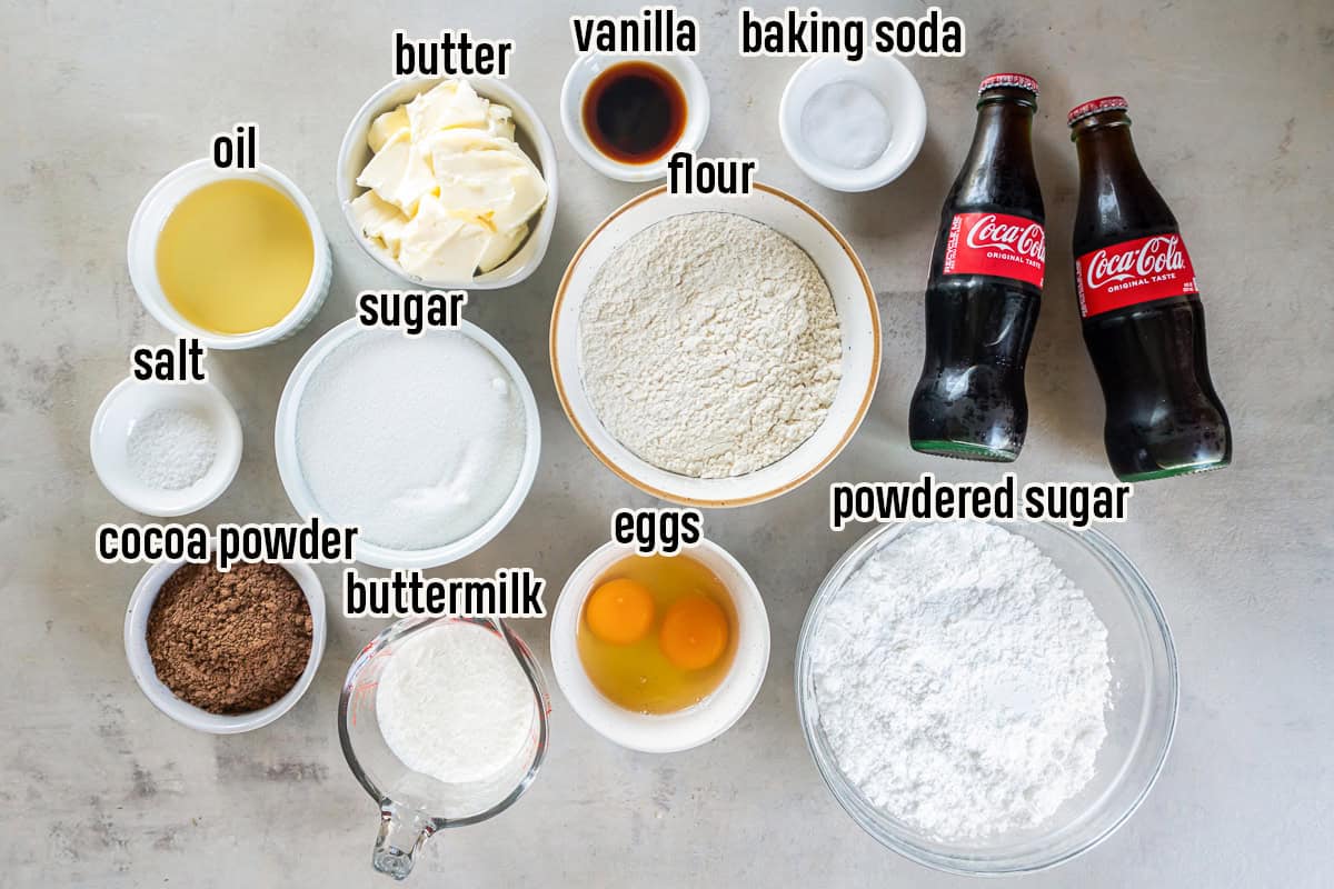 Ingredients for Coca-Cola Cake arranged on a countertop, including flour, granulated sugar, powdered sugar, cocoa powder, butter, vegetable oil, buttermilk, eggs, vanilla extract, baking soda, salt, and bottles of Coca-Cola.