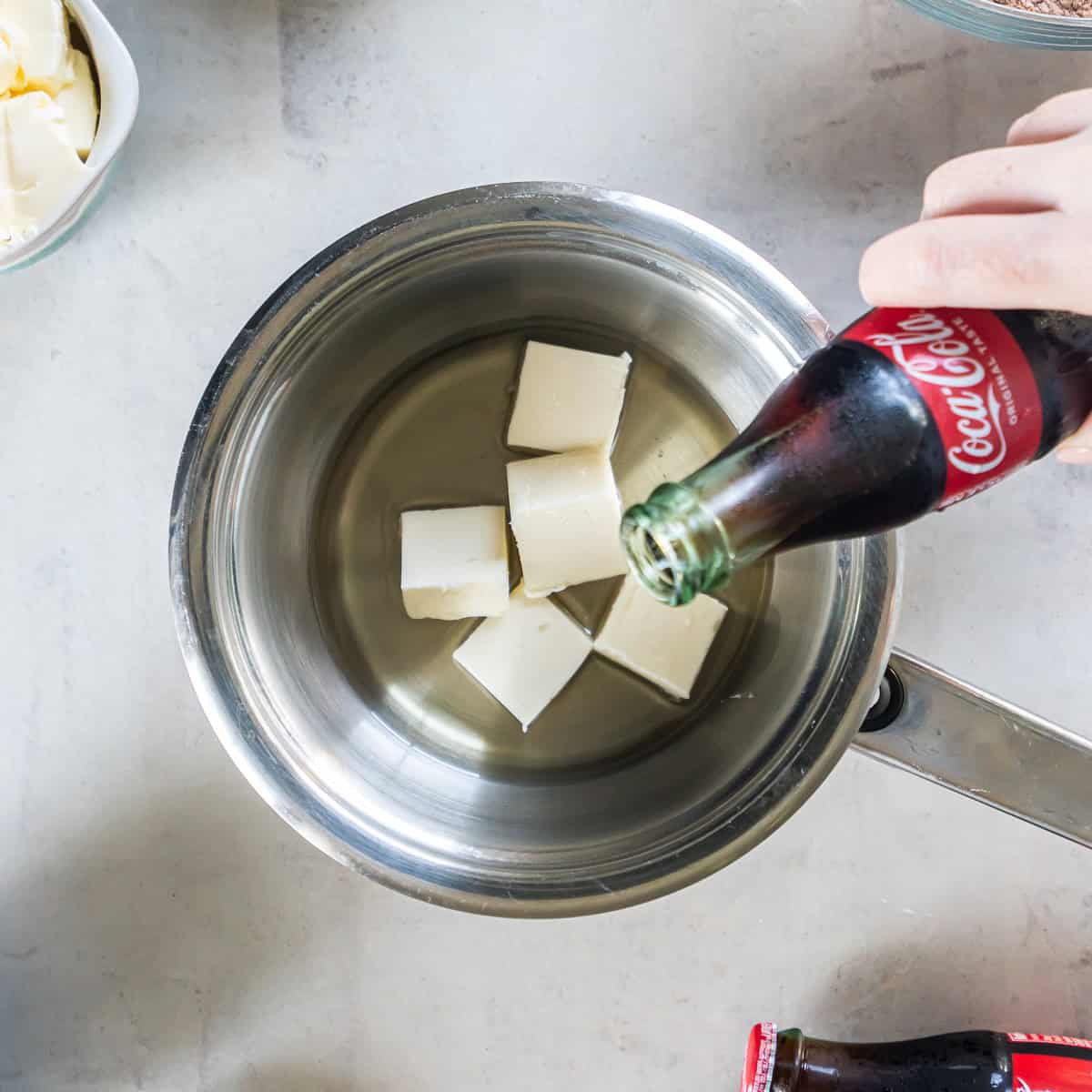 Coca-Cola being poured into a saucepan with butter and oil to prepare the cake batter.