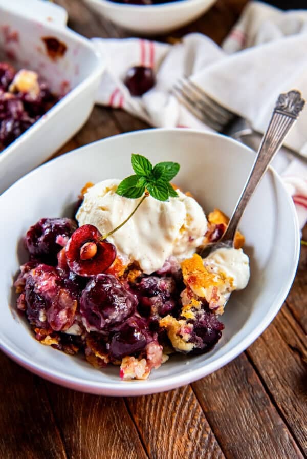 Fresh cherry cobbler topped with vanilla ice cream, a cherry half, and fresh mint in a white bowl.
