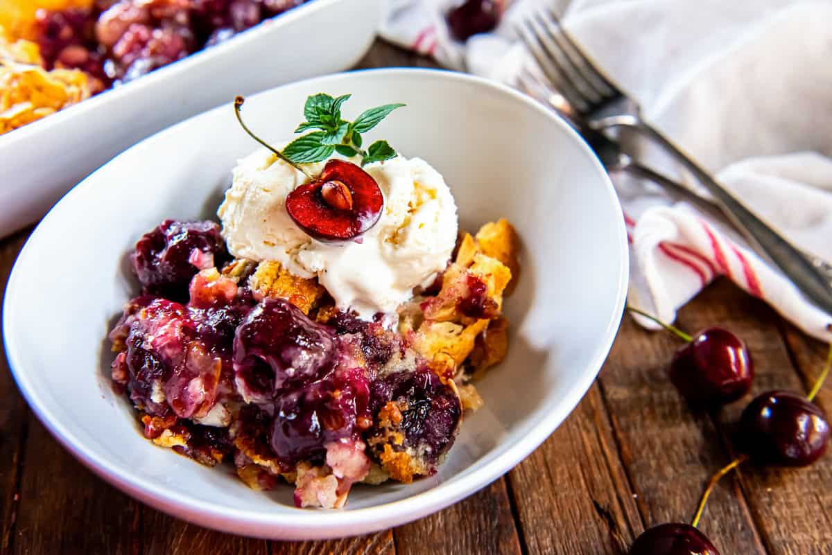 Fresh Cherry Cobbler topped with vanilla ice cream in a white bowl, served on a rustic wooden table with fresh cherries.
