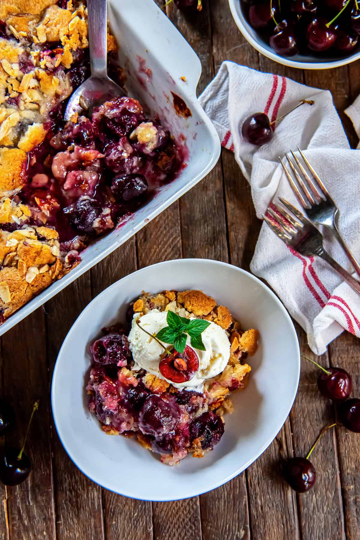 Fresh cherry cobbler topped with vanilla ice cream, a cherry half, and fresh mint, with the baking dish in the background.