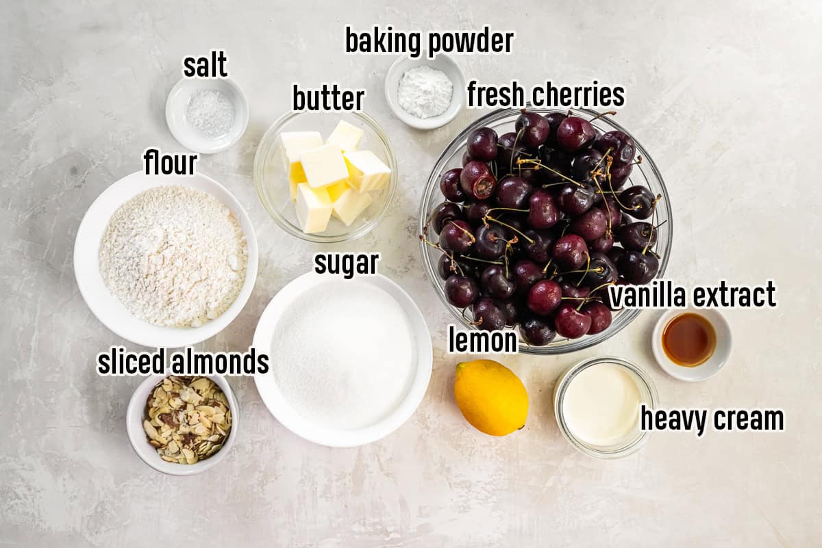 Ingredients for Fresh Cherry Cobbler arranged on a light surface, including fresh dark sweet cherries, flour, sugar, butter, heavy cream, lemon, vanilla extract, sliced almonds, baking powder, and salt.