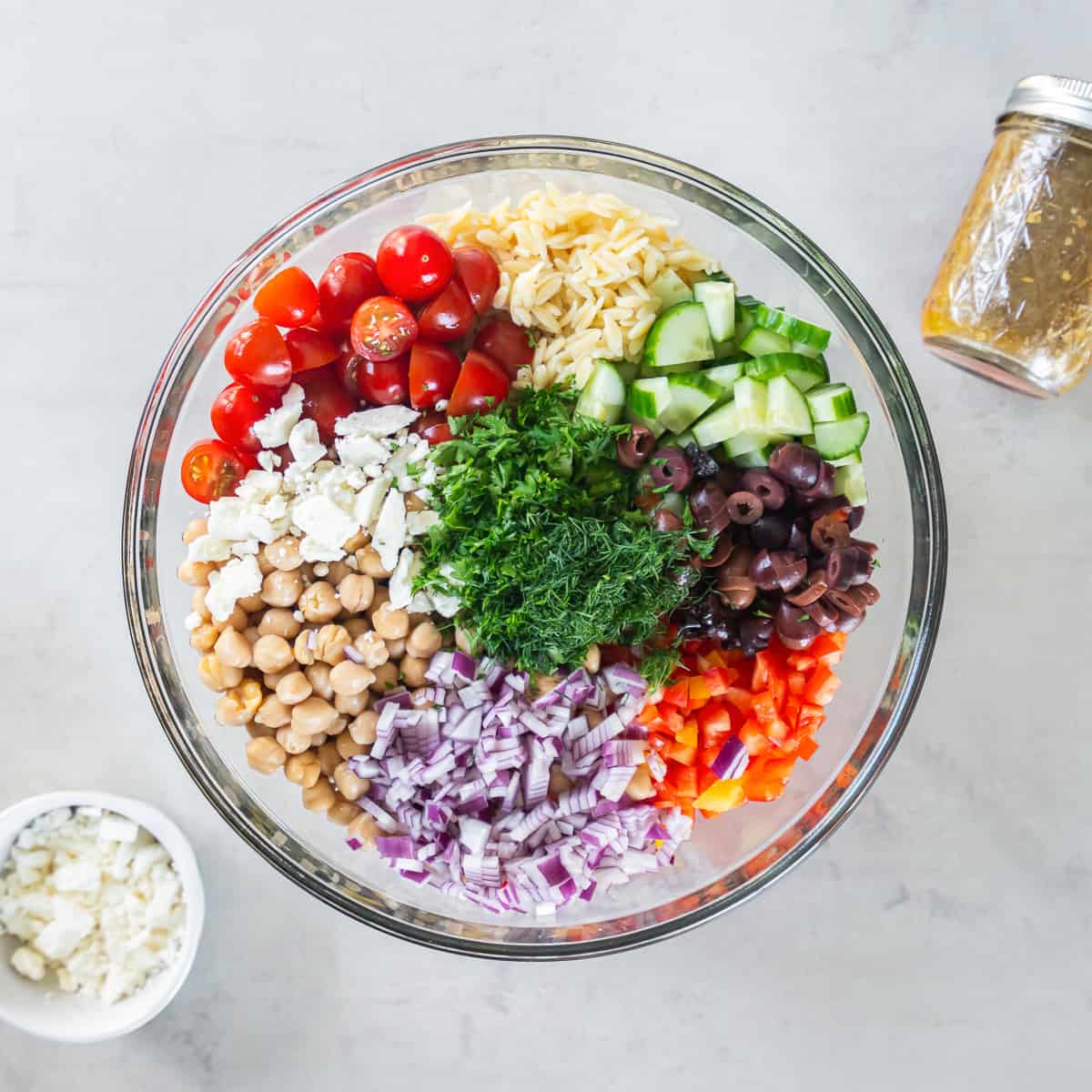 Greek Orzo Pasta Salad ingredients layered over cooked orzo in a large bowl before adding the vinaigrette and tossing to combine.