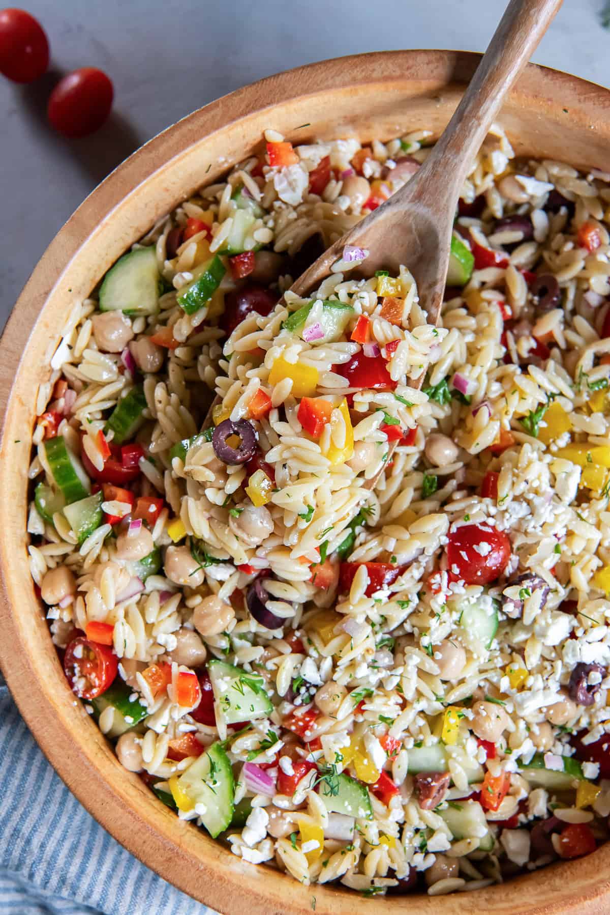 Wooden spoon lifting a serving of Greek orzo salad with chickpeas, cucumber, tomatoes, bell peppers, olives, feta, and fresh herbs.