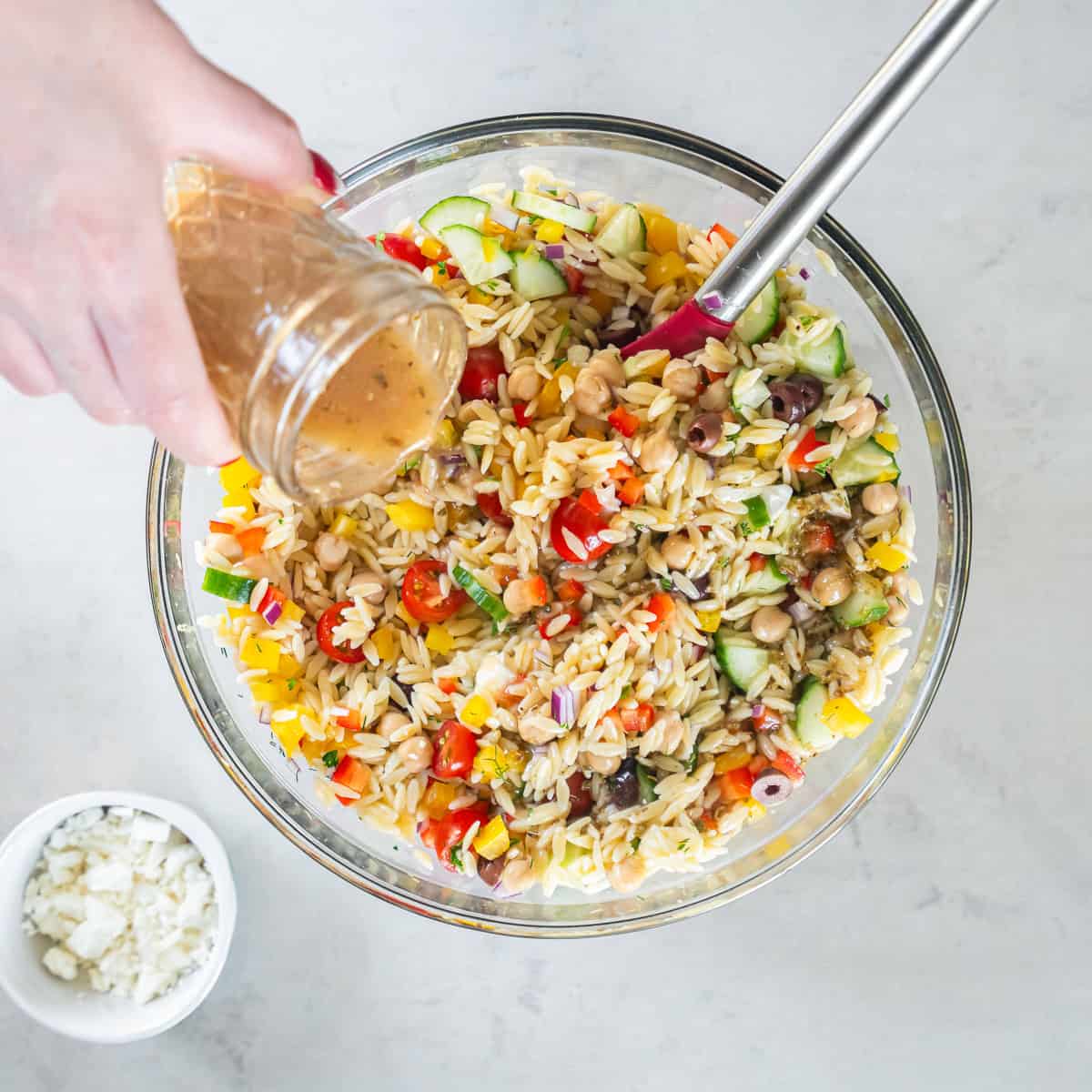 Pouring homemade Greek vinaigrette over Greek Orzo Pasta Salad with orzo, chickpeas, cucumber, bell peppers, cherry tomatoes, Kalamata olives, feta cheese, and fresh herbs before tossing.