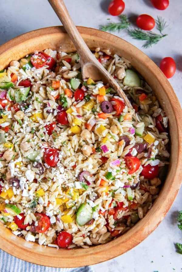Greek orzo salad with chickpeas, cucumber, tomatoes, bell peppers, olives, feta, and fresh herbs in a wooden serving bowl with a wooden serving spoon.