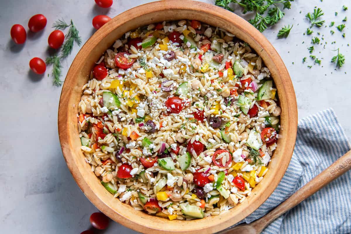 Greek orzo salad with chickpeas, cucumber, tomatoes, bell peppers, olives, feta, and fresh herbs in a wooden serving bowl.