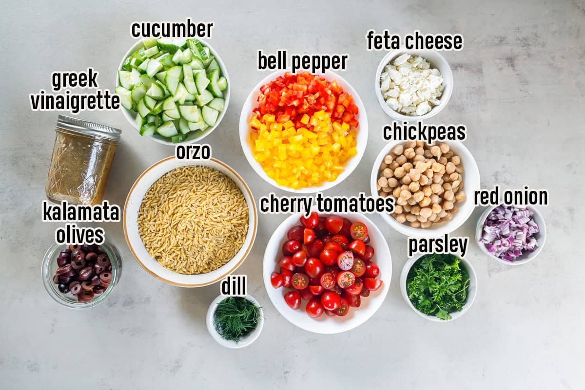 Ingredients for Greek Orzo Pasta Salad arranged in individual bowls, including cooked orzo, chickpeas, cucumber, cherry tomatoes, red and yellow bell peppers, red onion, Kalamata olives, feta cheese, parsley, dill, and homemade Greek vinaigrette.