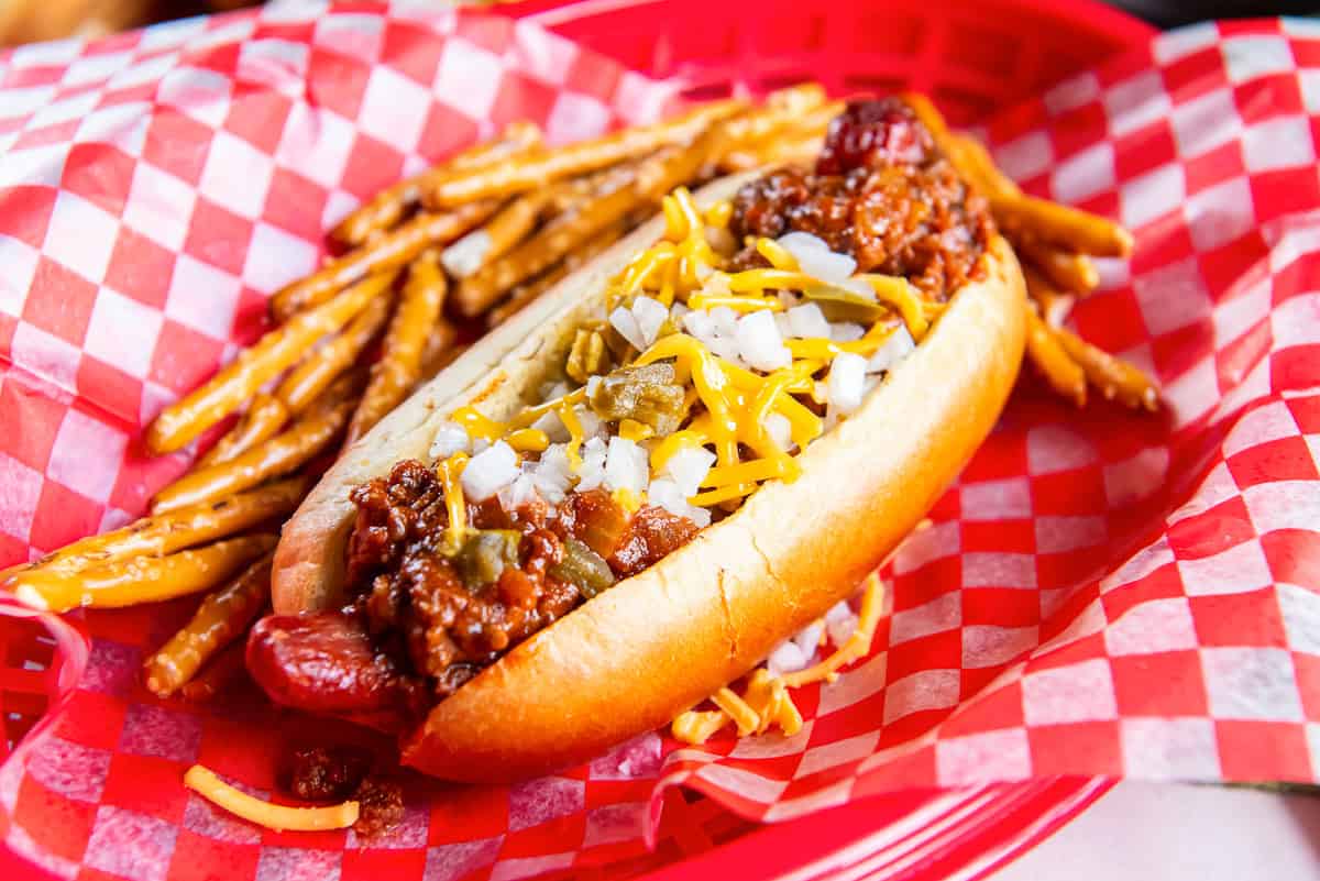 Chili dog topped with homemade hot dog chili, shredded cheddar cheese, diced onion, and chopped jalapeños served in a red basket with pretzel sticks.