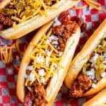 Three chili dogs topped with shredded cheddar cheese, diced onion, and chopped pickled jalapeños on red checkered paper.