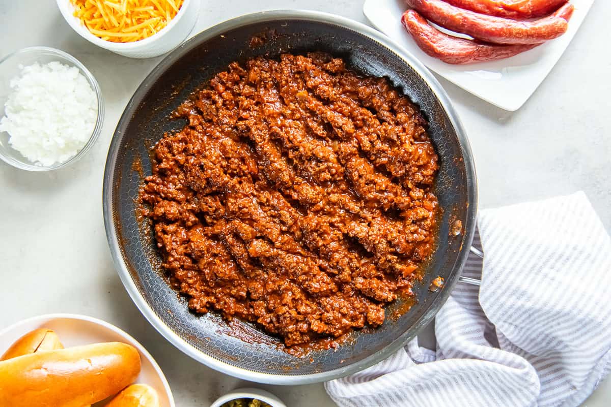 Homemade hot dog chili simmered in a skillet, with hot dog buns, shredded cheese, diced onion, pickled jalapeños, and hot dogs arranged around the pan for serving chili dogs.
