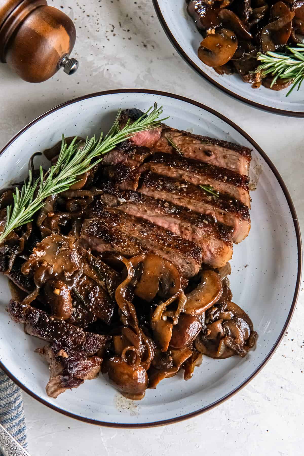 Sliced New York strip steak topped with sautéed mushrooms and onions on a white plate, garnished with fresh rosemary. A second plate and wooden pepper mills sit in the background.