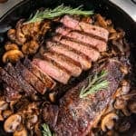 New York strip steaks with sautéed mushrooms and onions in a cast iron skillet, with one steak sliced to show a medium-rare center and garnished with fresh rosemary.